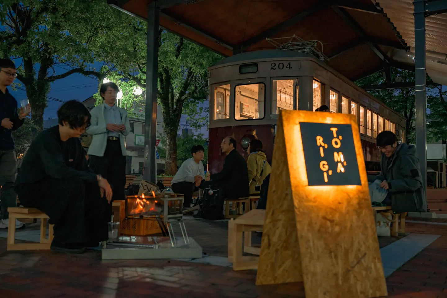 夜の駅前広場で焚き火を囲む人たちと、灯った TOMARIGI の行灯サイン