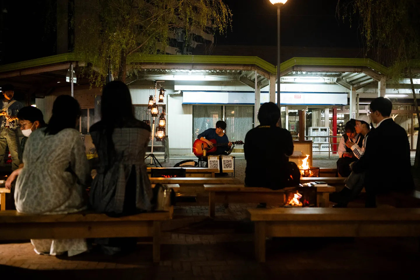 夜の広場のベンチに座る人たちと、灯ったランタン、奥でギターを弾く人
