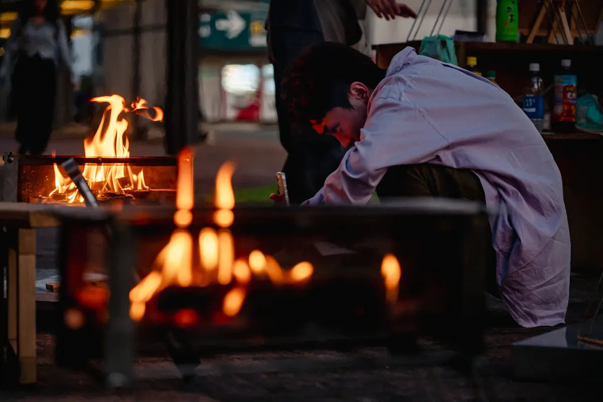 夜の駅前で、焚き火の炎ごしに一人でかがんで火の面倒を見ている人